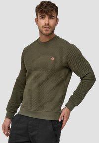 Sudadera verde oliva con un diseño texturizado y acolchado, cuello, puños y dobladillo acanalados; presenta un pequeño parche redondo con un logo en el pecho.