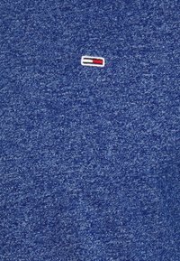 Petit logo rectangulaire avec des bandes blanches, rouges et bleu marine sur un fond de tissu texturé bleu.
