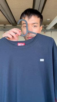 Άνδρας κρατάει μπλε σκούρο t-shirt της Diesel σε μαύρη κρεμάστρα κοντά στο πρόσωπό του, σε εσωτερικό χώρο με ορατές δοκούς οροφής.