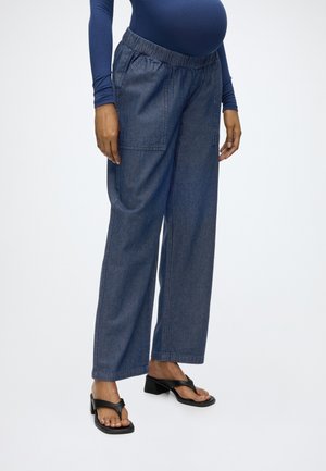 Gravid person iført mørkeblå langærmet top, løse mørke denimbukser med elastik i taljen og sorte sandaler med hæl, stående på hvid baggrund.
