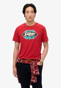 Camiseta de algodón roja con un estampado gráfico en negro y azul etiquetado como "Super Dry". Pantalones oscuros con una camisa de cuadros atada a la cintura. Cinturón marrón.