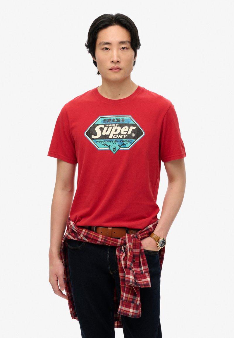 Camiseta de algodón roja con un estampado gráfico en negro y azul etiquetado como "Super Dry". Pantalones oscuros con una camisa de cuadros atada a la cintura. Cinturón marrón.