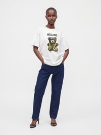 Γυναίκα που φοράει ένα λευκό φαρδύ T-shirt Moschino με γραφικό αρκουδάκι και σκούρο μπλε τζιν, στέκεται με τα χέρια στις τσέπες.