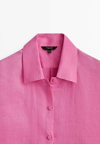 Chemise en lin rose avec col, équipée d'une fermeture boutonnée sur le devant et d'un tissu léger et texturé. Étiquette visible à l'intérieur du col.