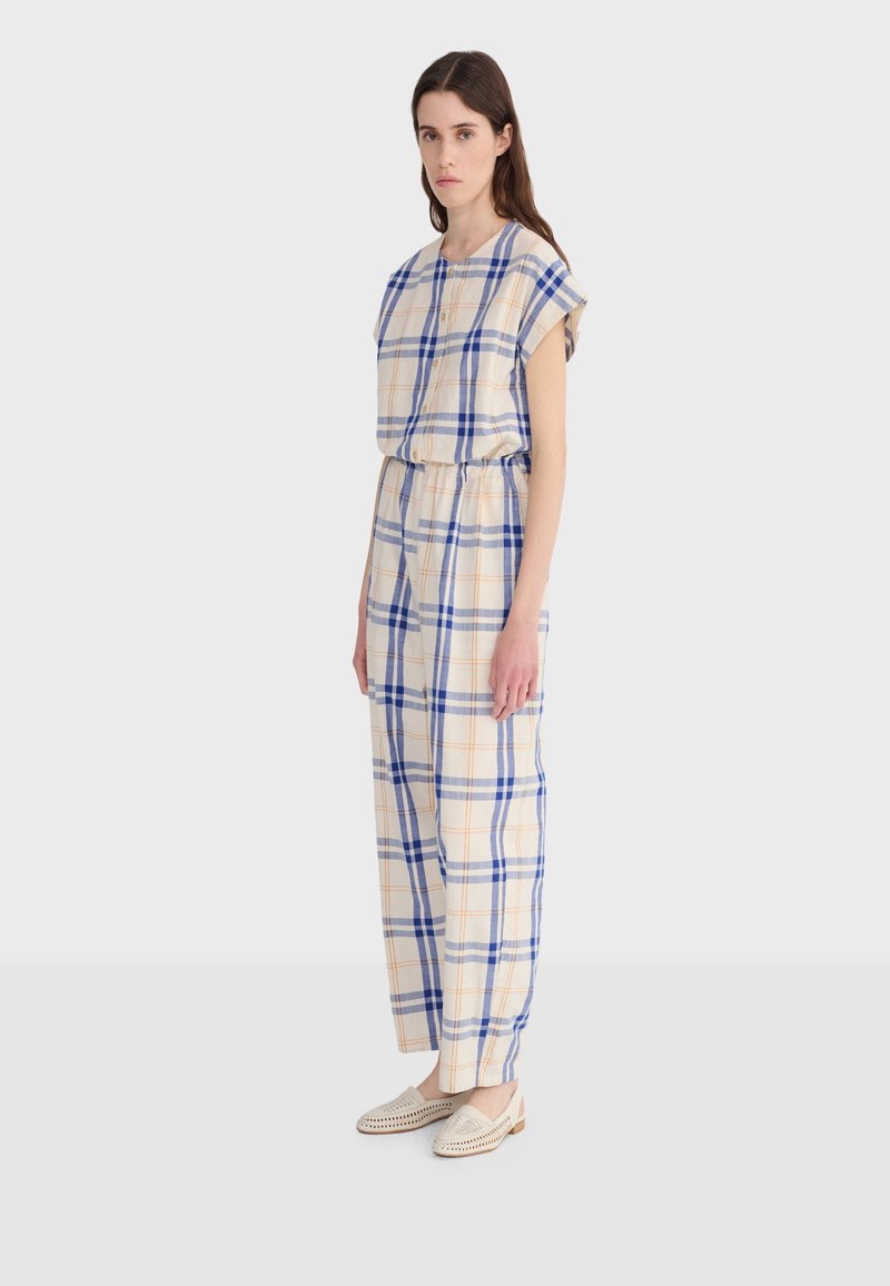 Femme debout portant une combinaison beige à carreaux avec des lignes bleues et orange, ainsi que des chaussures blanches tissées sans lacets, sur un fond clair.