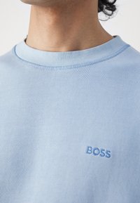 Man som bär en ljusblå tröja med rundad hals, broderad med "BOSS" på bröstet, vilket visar halsen och en del av käklinjen.