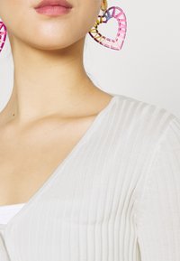 Bílý žebrovaný pletený top s výstřihem do V, doplněný velkými náušnicemi ve tvaru růžových a žlutých srdcí. Hladká textura, jednoduchý design.