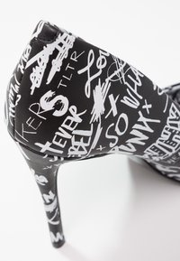 Chaussure à talons hauts noire avec une finition brillante, ornée de motifs graphiques manuscrits blancs. Bout pointu et talon aiguille élancé.