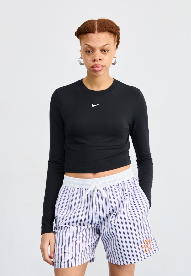 Svart langermet crop topp med hvit logo, paret med hvite shorts med lilla striper og oransje grafisk detaljer på siden.