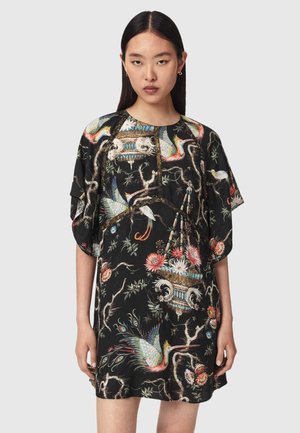 Femme aux cheveux raides et foncés portant une robe noire ample ornée d'imprimés colorés d'oiseaux, de branches et de fleurs, posant devant un fond uni.