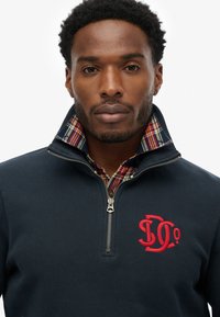 Felpa blu navy con mezza zip e logo ricamato rosso, con collo interno a motivo scozzese. Tessuto dalla texture liscia.