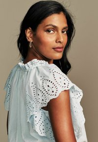 Blusa azzurra chiaro con ricamo a occhio di pernice, maniche a volant e scollatura ondulata. Tessuto morbido e dettagli intricati simili a pizzo.