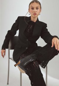 Svart skräddarsydd kavaj med en slät yta, bärs över en svart spetsklänning. Har två knappar och sitter i en position på en metallstol.