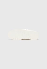 Semelle en caoutchouc blanche avec un motif texturé et de légères rainures, présentant un détail de logo noir. Fini lisse avec un léger éclat.