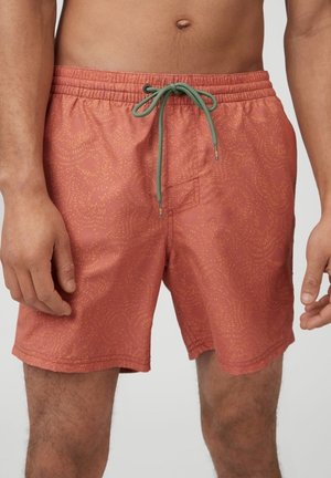 Primer plano de una persona que lleva pantalones cortos de baño color coral con un sutil patrón amarillo y un cordón verde atado en la cintura.