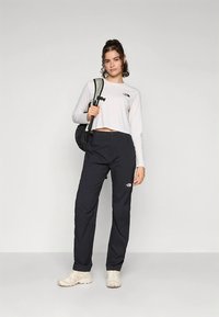 Bela obrezana majica z dolgimi rokavi z logotipom, črne lahke hlače in bež čevlji. Črni nahrbtnik z obeskom za teniško lopar na ramenu.