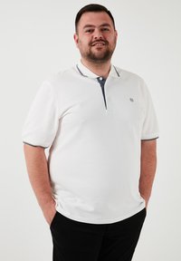 Biela polo košeľa s tmavo modrým golierom, krátkymi rukávmi a tromi gombíkmi. Na hrudi sa nachádza malé logo a látka má textúrovaný vzor. Čierne nohavice.
