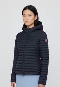 Navy quilted puffer jacket with a hood, featuring horizontal stitching, zip closure, and a logo patch on the sleeve. Lightweight fabric.