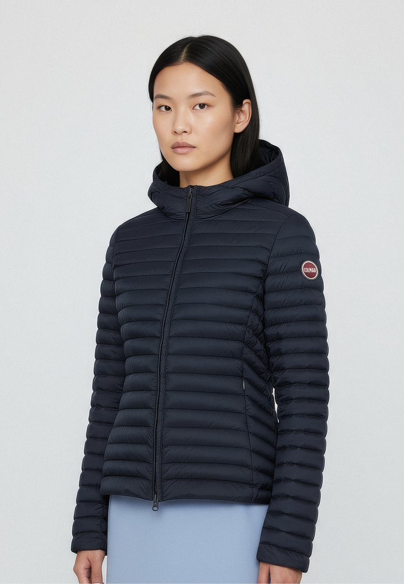 Navy quilted puffer jacket with a hood, featuring horizontal stitching, zip closure, and a logo patch on the sleeve. Lightweight fabric.