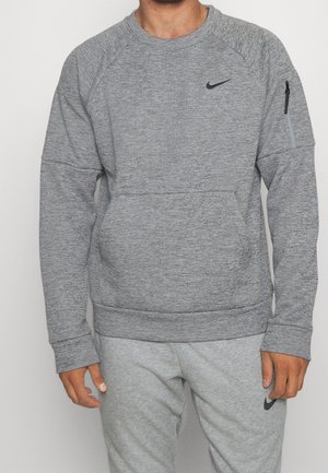 Mężczyzna w szarej bluzie Nike z kieszenią na suwak na rękawie i dopasowanych szarych spodniach dresowych Nike, stojący na jednolitym tle.