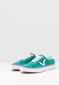 Turkosa mockasneakers med vita snören, med en svartvit logotyp på tungan, vit gummisula och kontrasterande detaljer med logotyp på sidan.