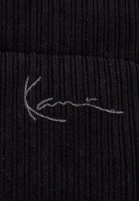 Mustast fliisist riie, millel on vertikaalne soonimine ja valge tikandiga allkiri. Tekstuur tundub pehme ja veidi udune.