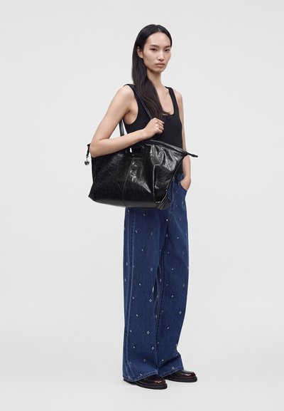 Femme portant un débardeur noir et un pantalon large bleu, tenant un grand sac cabas en cuir noir sur son épaule devant un fond uni.