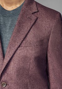Blazer bordeaux en tissu texturé, avec des revers crantés, une poche poitrine et une fermeture à un seul bouton. Pull gris à col rond en dessous.
