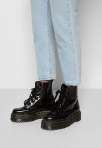 Botines negros de charol con suela de goma gruesa, cordones en la parte delantera y detalles de costura visibles. Combinados con unos vaqueros de un azul claro.
