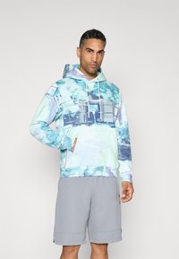 Felpa con cappuccio in blu chiaro e teal con motivi astratti ad acquerello, tasca a marsupio e cordini bianchi. Abbinata a pantaloni corti grigi.