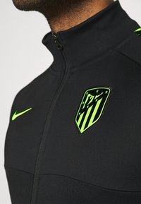 Chaqueta negra con cremallera y cuello alto. Presenta un brillante logo de Nike en verde y un emblema del club con rayas y estrellas. Tejido liso y atlético.