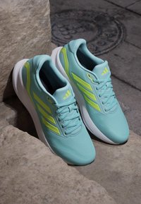 Baskets de sport bleu clair avec des accents jaunes, tige en maille, tissu texturé et semelle intermédiaire blanche, placées sur une surface en pierre.