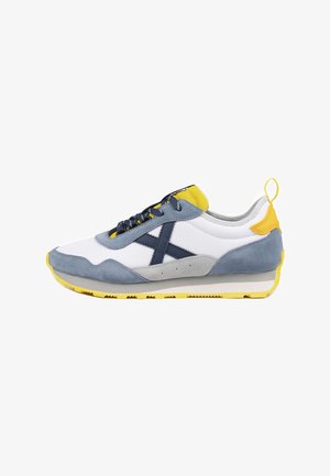 Zapatilla deportiva con cuerpo de malla blanca, detalles en ante gris y azul, contrafuerte trasero amarillo y suela de goma texturizada con un patrón de dibujo visible.