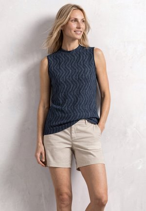 Lyshåret kvinde iført en ærmeløs marineblå mønstret top og beige shorts står op ad en lys væg, smilende og kigger til siden.