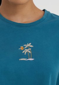 Turqouise katoenen T-shirt met een geborduurd ontwerp van twee palmbomen en een zon op een lichtroze achtergrond, wat een strandachtige scène creëert.