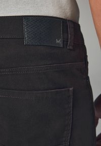 Dark brown pants with a textured black leather waistband. Features visible stitching, pockets, and a woven pattern on the leather accent.