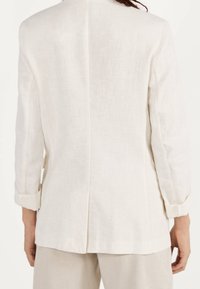 Bílý lněný blejzr s jednoduchým designem, dlouhými rukávy a jediným rozventem vzadu. Tkanina má přirozenou texturu a je lehká.