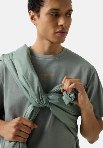 Mladý muž hledící stranou, oblečený v šedém tričku s nápisem "FIRE+ICE", který si kolem ramen uvazuje zelenou bundu.