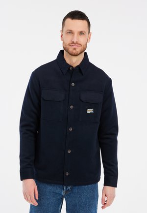 Marineblauwe fleecedonker jacket met een kraag, twee borstzakken, knoopsluiting en een klein logo-patch. Lange mouwen met knoopmanchetten.