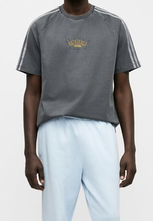 Person trägt ein dunkelgraues Adidas Originals Kurzarmshirt mit weißen Streifen an den Ärmeln und hellblauen Hosen vor einem einfarbigen Hintergrund.