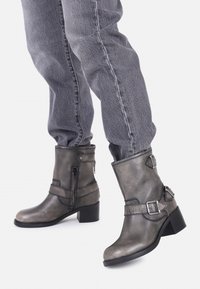 Botas de piel gris hasta el tobillo con un acabado ligeramente brillante, tacones negros más gruesos, cremallera lateral y correas decorativas con hebillas.