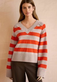 V-hals sweater med brede orange og grå striber, ribbede manchetter og nederste kant. Blødt materiale med en afslappet pasform, parret med mørke bukser.