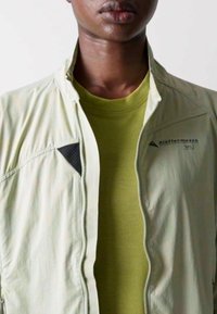 Veste légère vert clair avec fermeture éclair, présentant une chemise verte texturée en dessous. Accents noirs notables sur la veste.