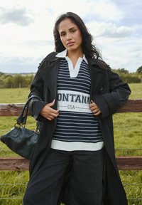 Cappotto lungo nero su una polo a righe blu navy e bianche con la scritta "MONTANA USA". Sono visibili pantaloni scuri e una borsa nera con texture.