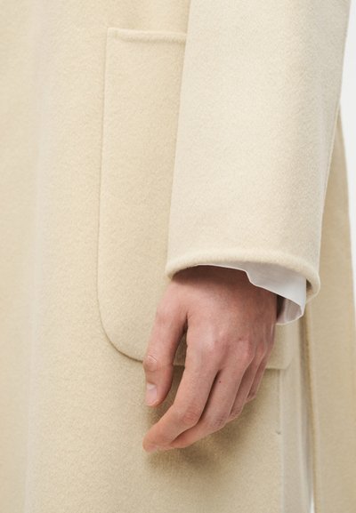 Ανδρικό χέρι δίπλα στην τσέπη ενός μπεζ παλτού από μαλλί με μανσέτα λευκού πουκαμίσου να φαίνεται στον καρπό.