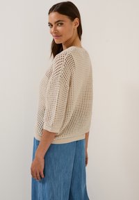 Beige gestrickter Pullover mit lockerem, offenem Gewebe, Dreiviertelärmeln und einem entspannten Schnitt. Kombiniert mit einem hellblauen Jeansrock.