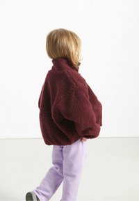 Bordo flis jakna s visokim ovratnikom, oversized kroja i teksturiranom površinom, u kombinaciji sa svijetloplavim hlačama.
