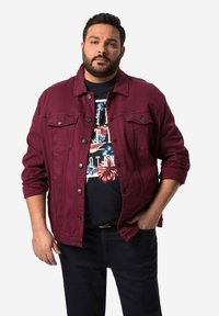 Bordový denimový kabát s knoflíkovým zapínáním a náprsními kapsami, přehozený přes černé tričko s potiskem obsahujícím květinové vzory, doplněný o tmavé kalhoty.