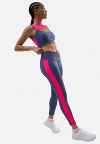 Conjunto deportivo que incluye una parte superior corta de color azul oscuro con acentos en rosa y corte, combinado con legging azul oscuro con paneles laterales rosas. Zapatillas blancas.