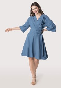 Robe portefeuille bleue en tissu doux, avec un décolleté en V, des manches larges, une taille nouée avec une quincaillerie, et une jupe évasée.
